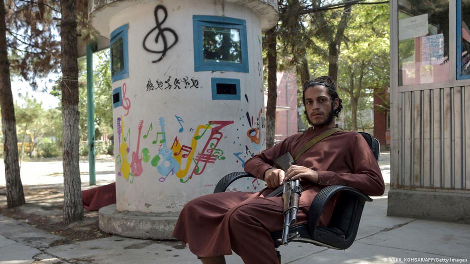 An armed Taliban fighter lounges in a chair at the entrance to the Afghanistan National Institute of Music compound in September 2021; a treble clef graffito can be seen on the pillar behind him