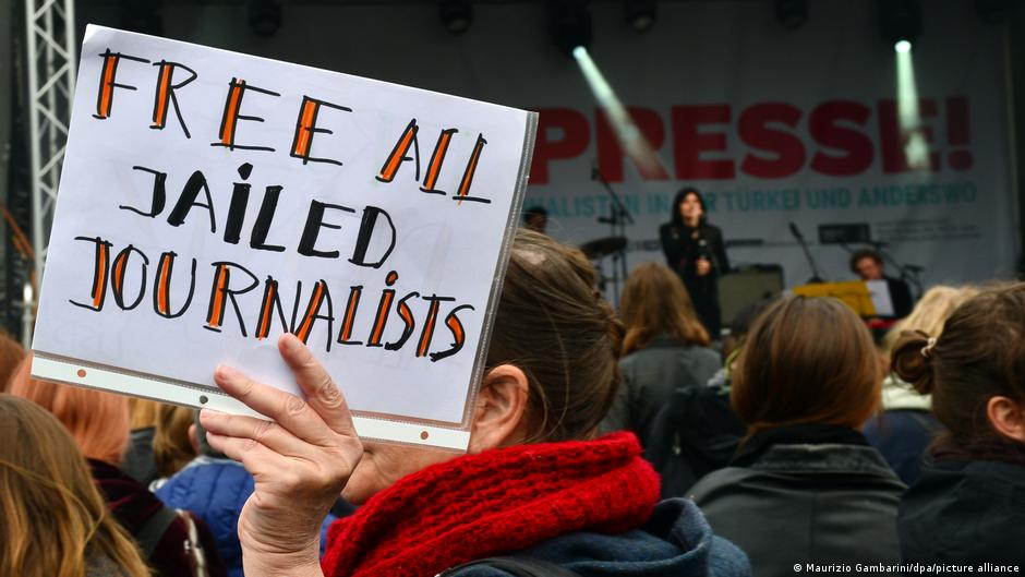 A woman in a crowd holds up a sign in front of her face that reads "Free all jailed journalists"