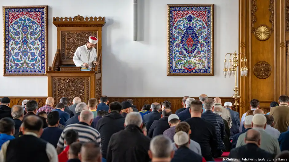 An imam is seen standing at a podium in front of a group of Muslims during Friday prayers in the Merkez DITIB Mosque in Duisburg, Germany, there are decorative images on the wall