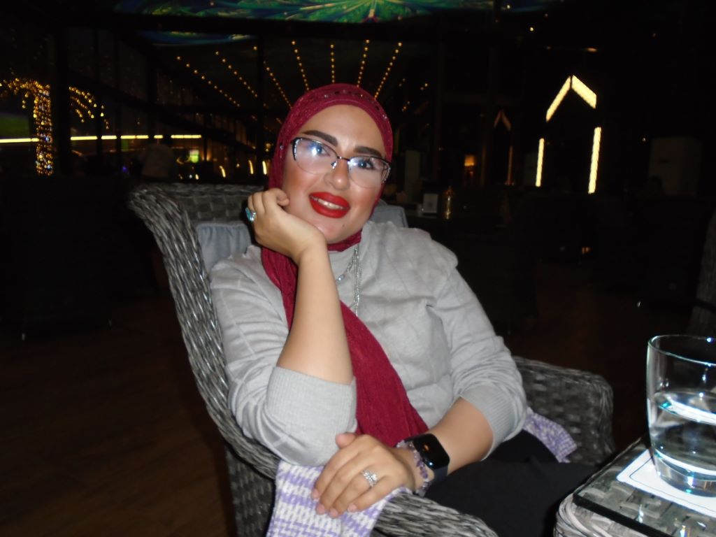 Rawan Al Zaidi, dressed in pale grey with a red headscarf sits in a grey wickerwork chair and smiles