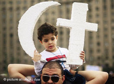 Egyptian protester carrying a child, holding a crescent and a cross made of polystyrene (photo: picture alliance/dpa)