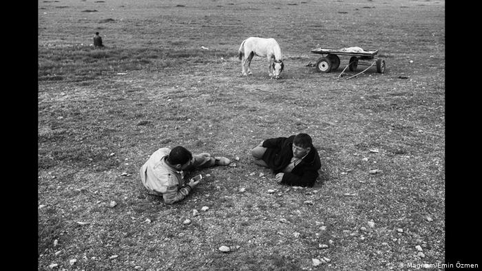 A balck-and-white picture shows two men lying on the ground, with one checking his phone while horse and cart stand in the background (photo: Magnum/Emin Ozmen)
