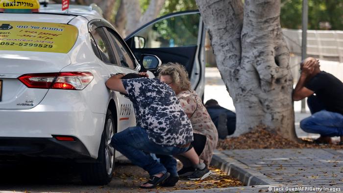 Three people duck behind a car and under a tree, covering their heads with their hands