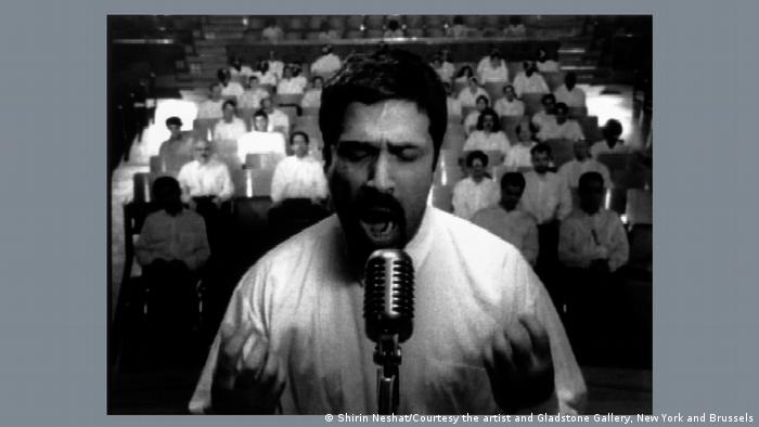 Das schwarz-weiße Bild eines Mannes, der ein weißes Hemd trägt und passioniert in ein Mikrofon spricht. Dahinter sitzen weitere Männer in weißen Hemden. Ausschnitt aus einer Videoinstallation.