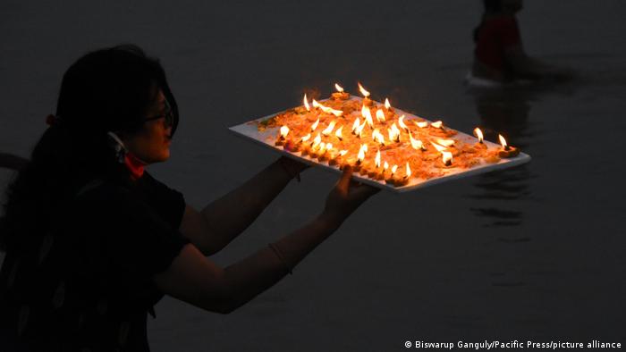Eine Frau hält ein Tablett mit angezündeten Kerzen und Blüten in den Händen und steht vor dem Fluss Ganges. Es ist dunkel.