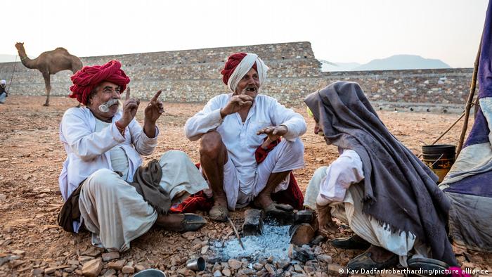Drei Männer, die in Tücher gekleidet sind, sitzen zusammen vor einem gelöschten Feuer und gestikulieren und lachen. Im Hintergrund sind eine Mauer, ein Kamel und Berge zu sehen.