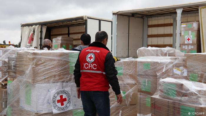 A man in a Red Cross waistcoat stands in front of packages shrink-wrapped in foil.