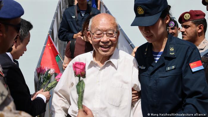 A Chinese man exits a ship via a gangway and is met at its end by uniformed people. Someone holds out a pink rose to him.
