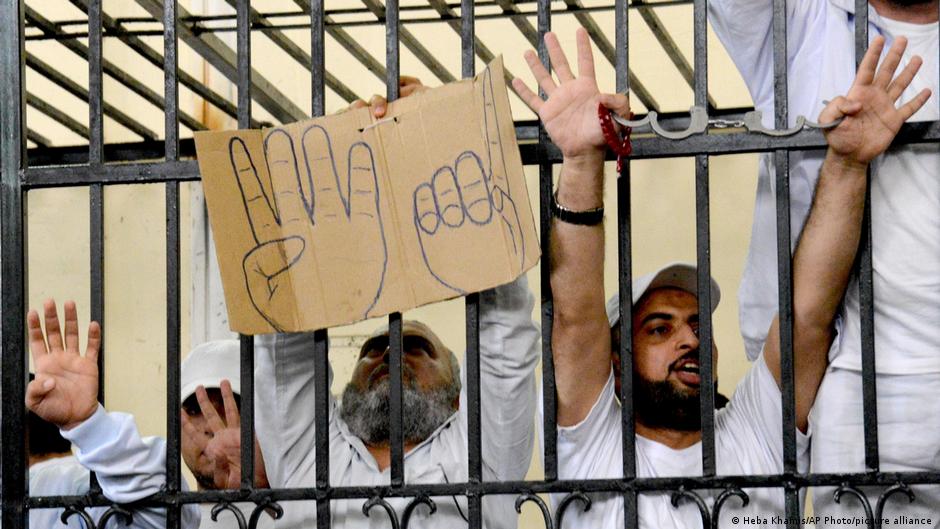 أشخاص في قفص الاتهام يرفعون بأصابعهم شعار احتجاجات رابعة العدوية - مصر. Defendants in the Rabaa trial hold up the Rabaa symbol and wave from the defendants' cage (image: Heba Khamis/AP Photo/picture alliance) 
