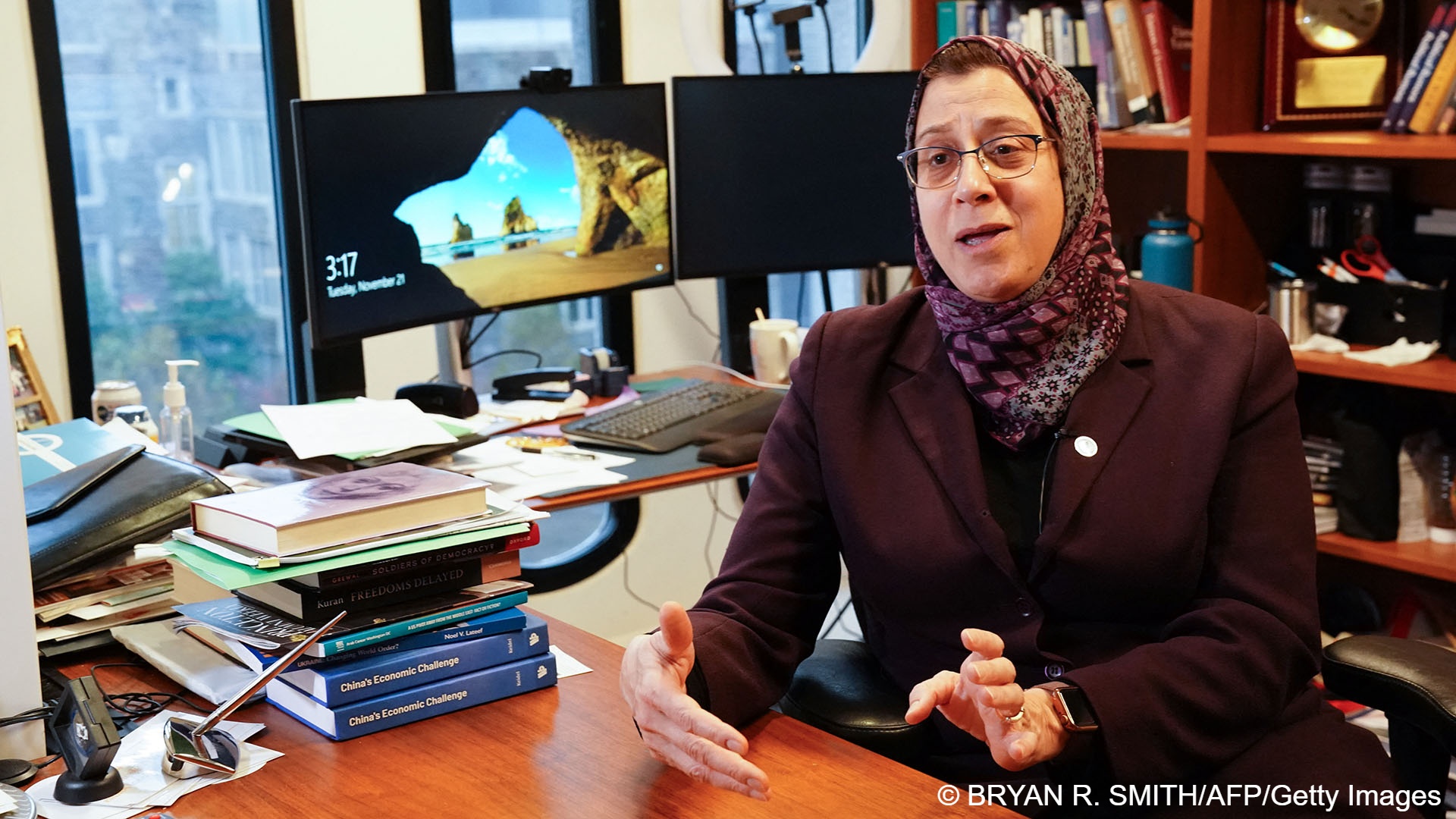 Wearing a headscarf, Amaney Jamal sits at her desk in an office strewn with books and papers. In the background are two monitors and book shelves
