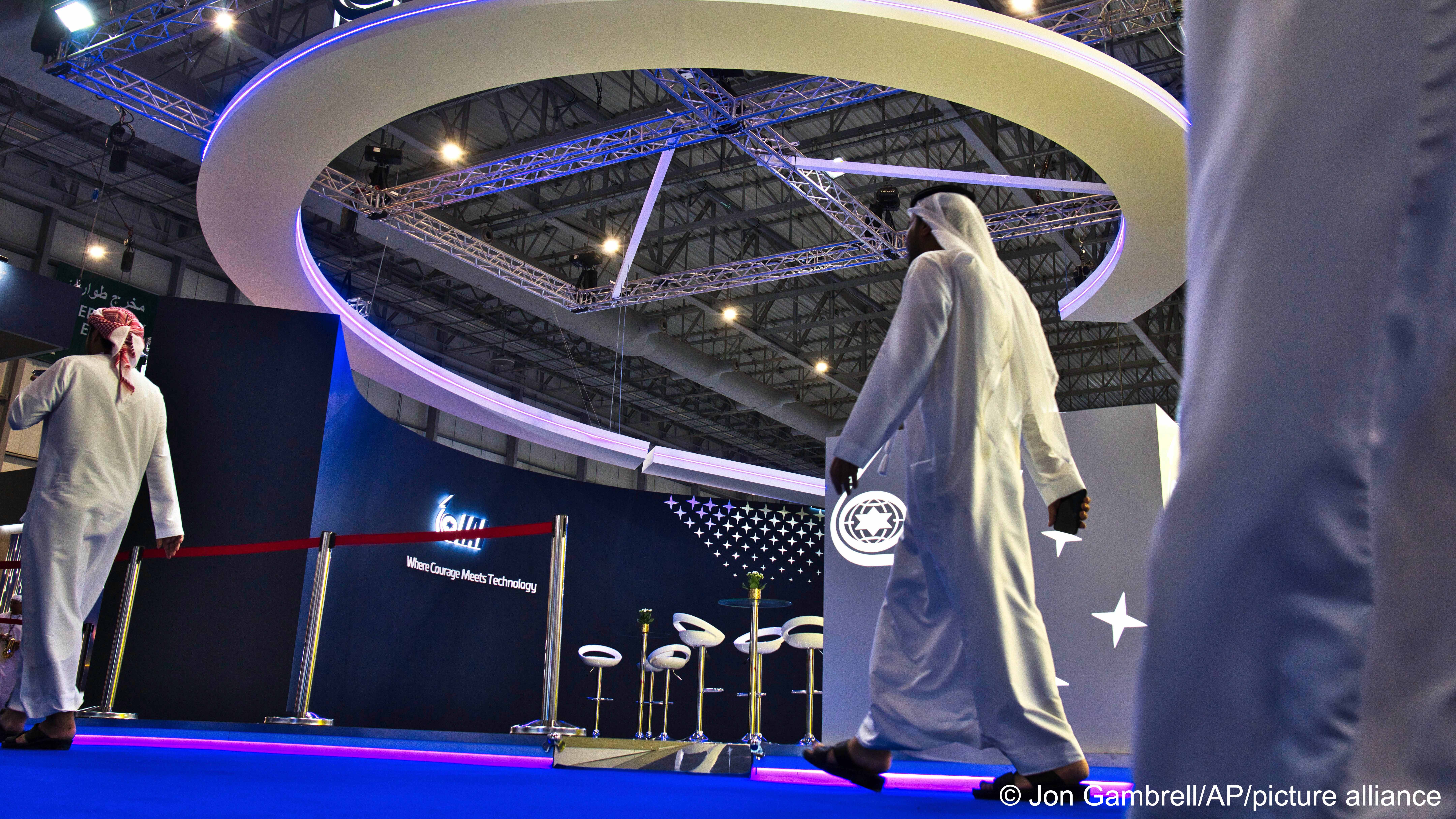 Men in Arab dress walk past an empty dark blue exhibition stand with a curved white ceiling element