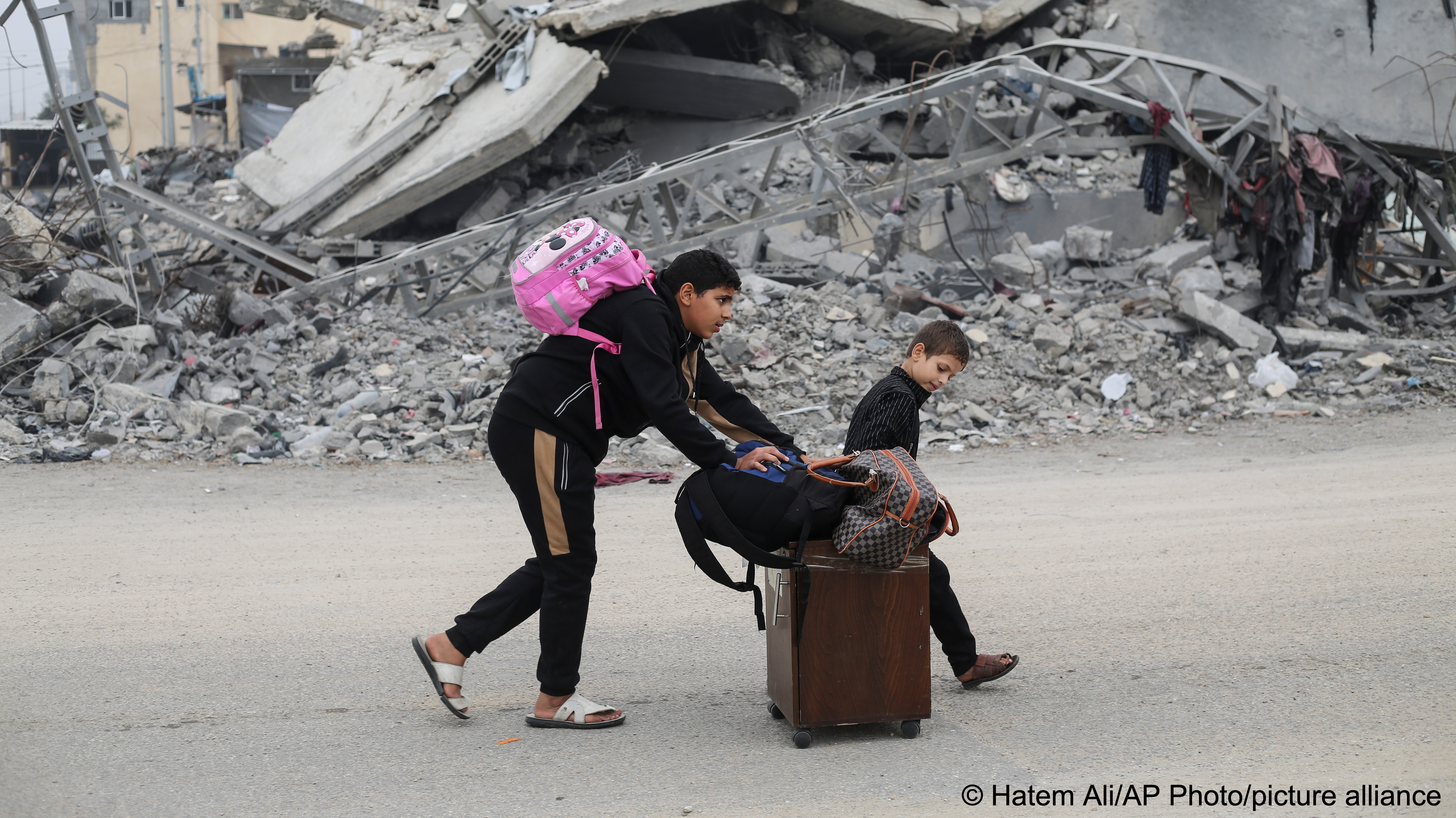 Two young Palestinians push a wooden cabinet on wheels and several bags along a road. In the background are piles of rubble from destroyed buildings, Rafah, Gaza Strip, 5 December 2023