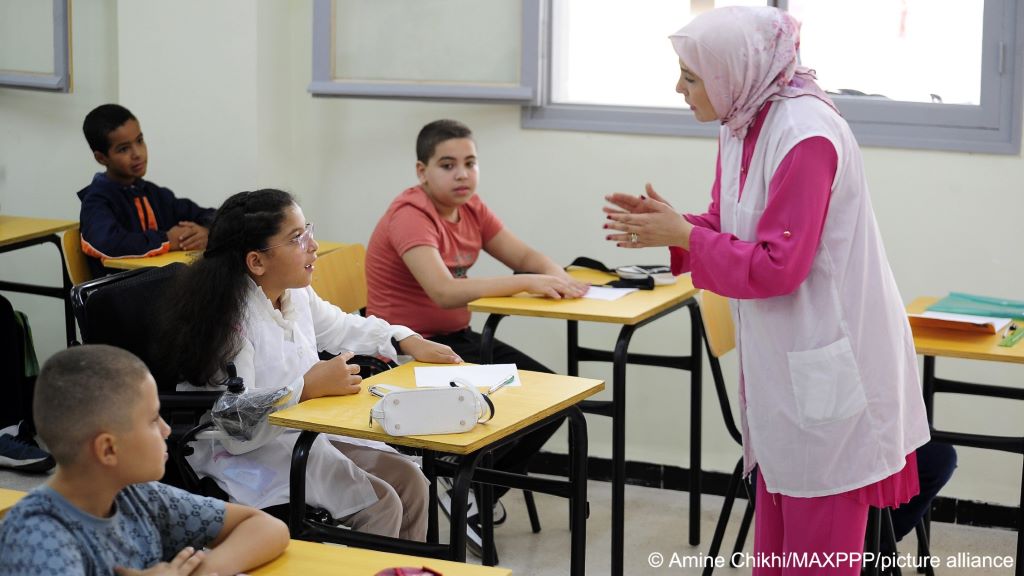An Algerian primary school teacher dressed in pink and white gestures as she speaks to a girl in her class; other pupils can be seen at other tables in the classroom