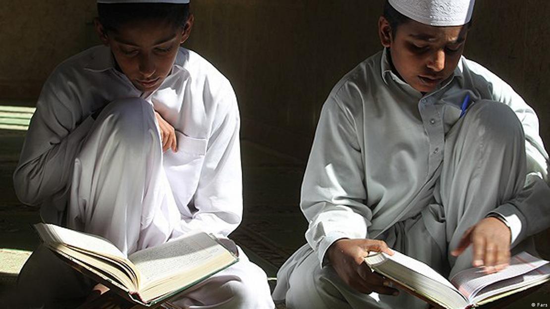 Two Iranian Sunni boys dressed in white sit side by side on the floor reading the Koran