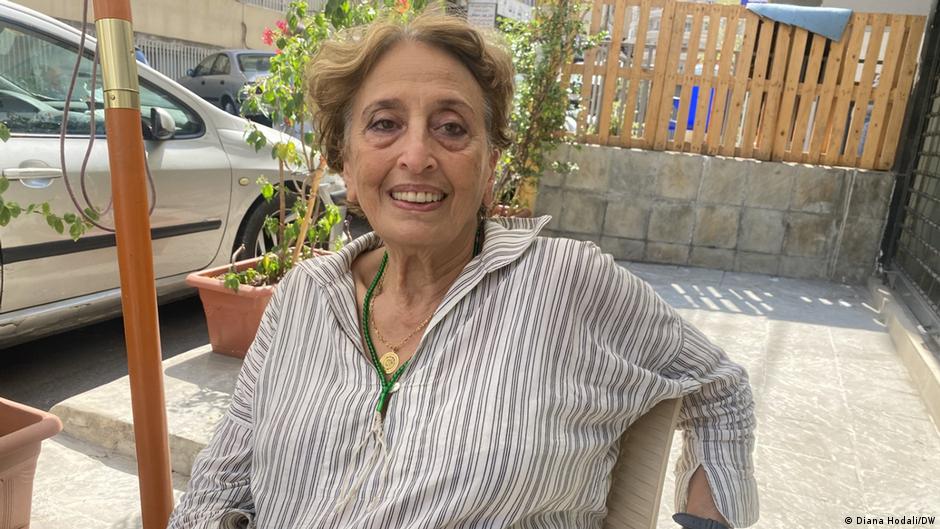 Lebanese entrepreneur Nawal Traboulsi sits in a chair outside her shop in Beirut. There is a car parked on the left and a slatted fence behind her
