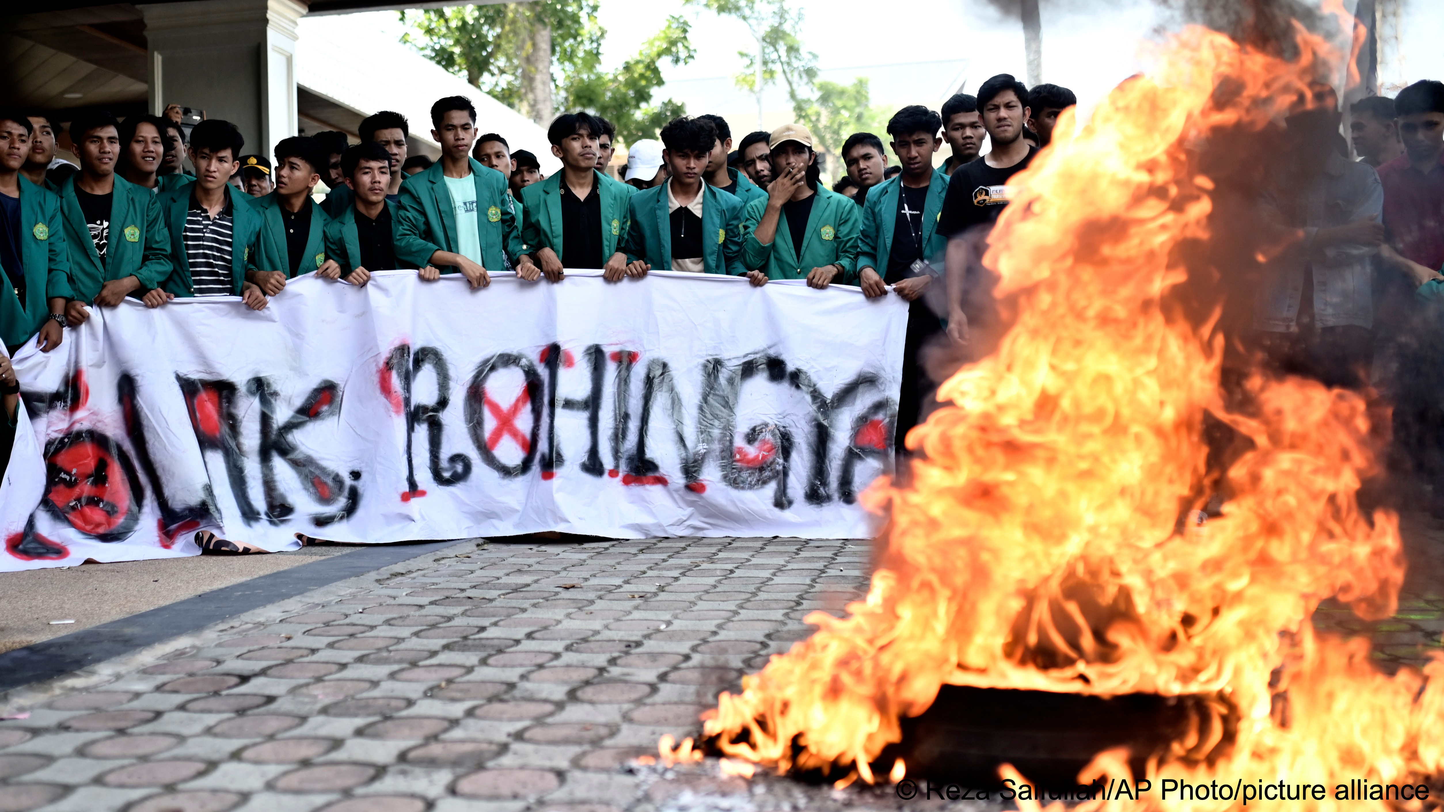 A group of students dressed in green tops and holding a large banner watch as tyres burn during a protest against Rohingya refugees in Banda Aceh, Indonesia, 27 December2023
