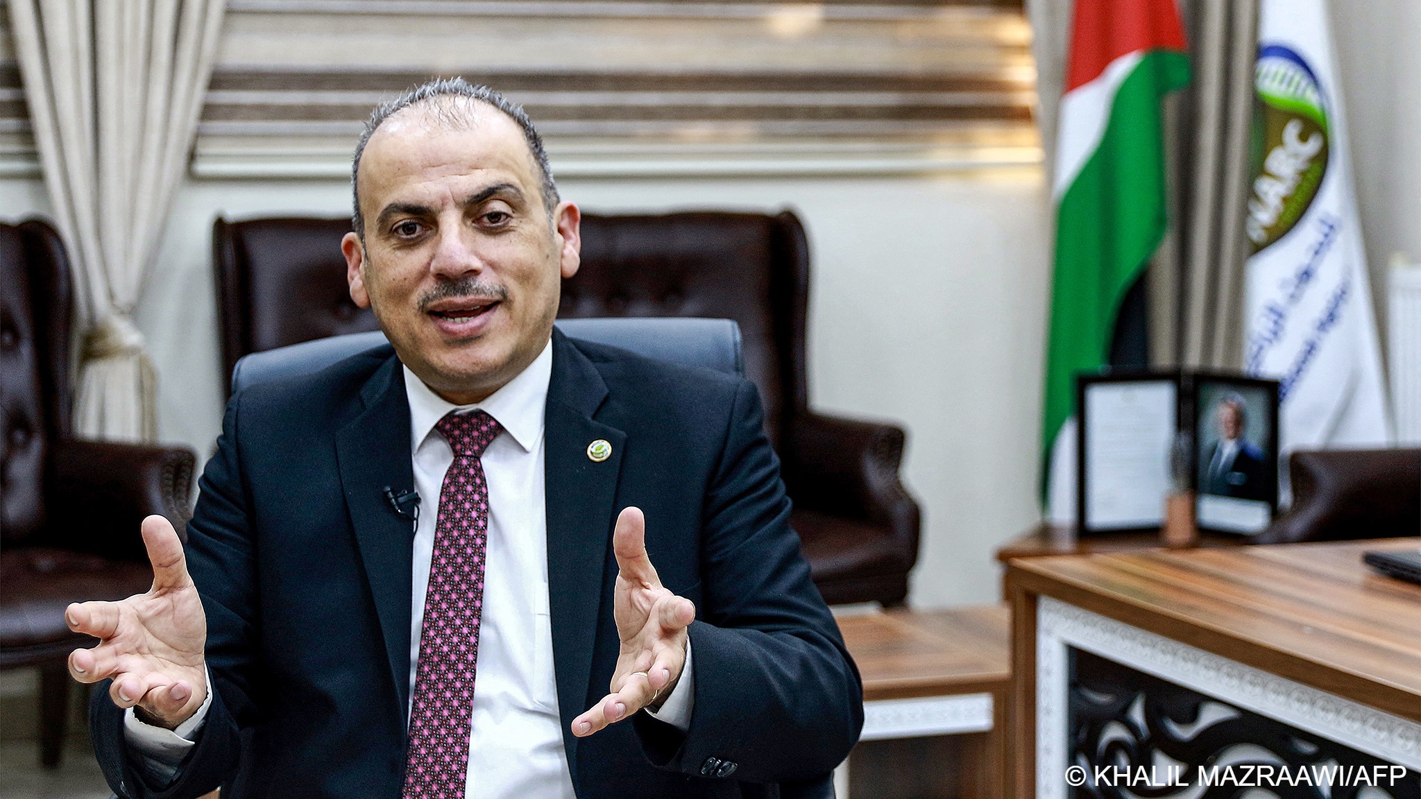 Nizar Haddad, director of the Jordanian National Agricultural Research Center, gestures as he speaks, sitting on a chair with the flags of Jordan and the NARC behind him