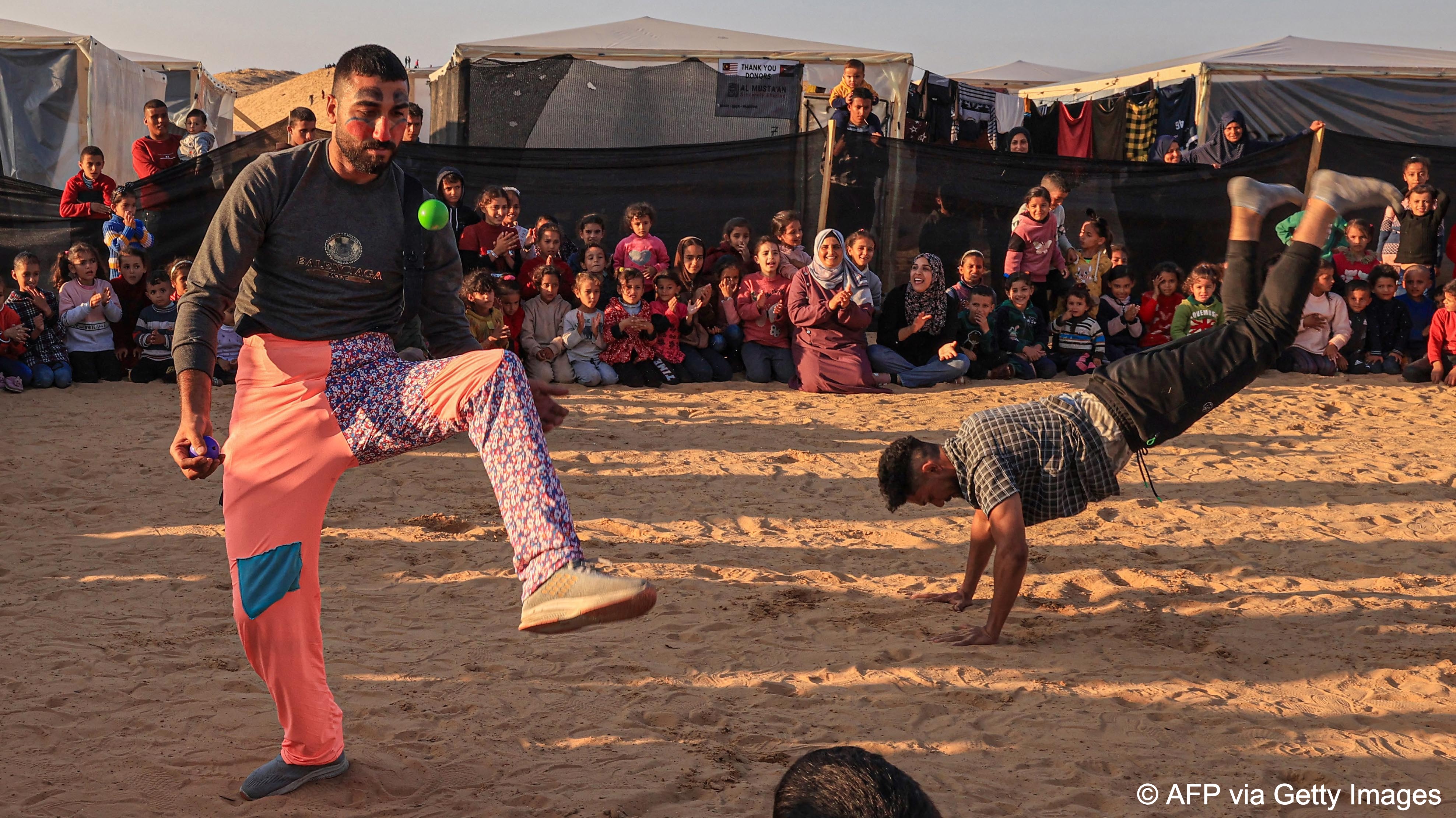 One man juggles and another man does acrobatic tricks to entertain a crowd in a camp near Rafah