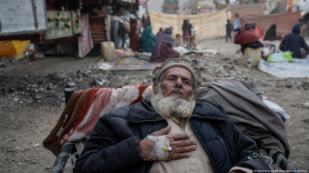 An elderly Afghan man receives medical care. He is leans back on a chair covered in blankets. In the background are trucks and people sitting on mats on the ground. 