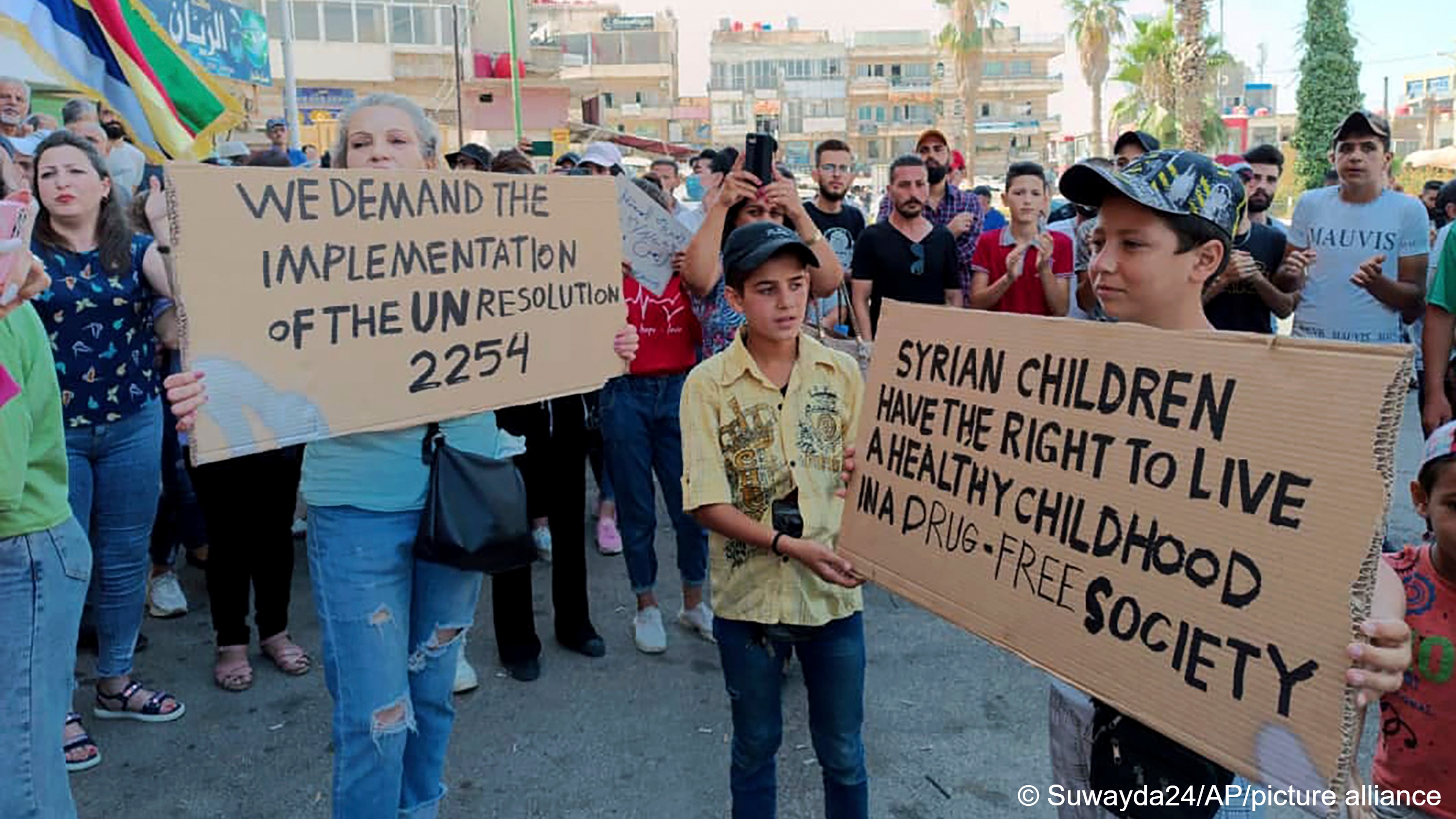 A woman and children hold cardboard placards demanding the implementation of UN Resolution 2254 and a drug-free society. In the background is a crowd of people, some clapping, some holding up Druze flags, Sweida, Syria, 26 August 2023