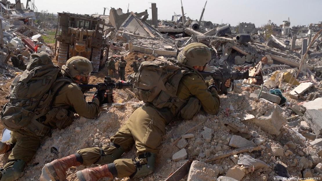 Two Israeli soldiers in full combat gear lean on a pile of rubble and train their weapons on a target in the distance. They are surrounded by rubble and destroyed buildings, Gaza Strip, February 2024