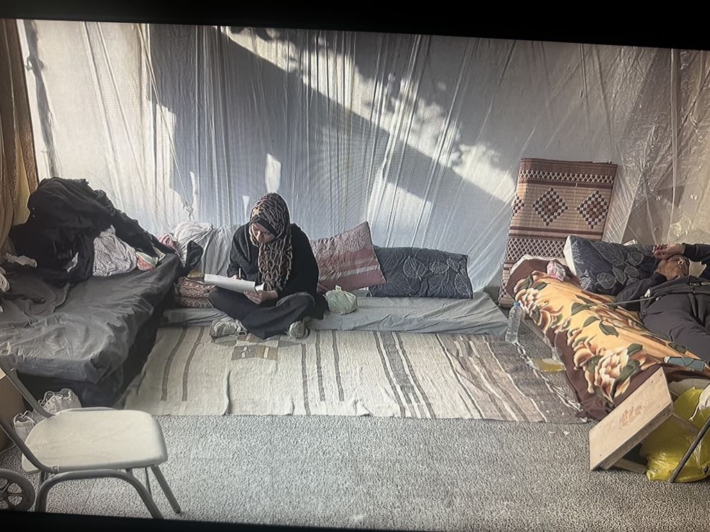 A woman in an animal-print headscarf and black clothes (Menna Hamouda) sits cross legged on the floor of a sparsely furnished tent and draws; on the right, a person lies on a mattress on the ground