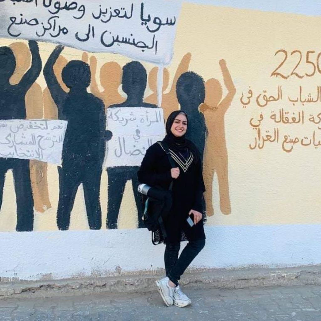 A young woman dressed mostly in black and wearing a headscarf (Menna Hamouda) smiles broadly as she stands in a relaxed pose in front of a mural