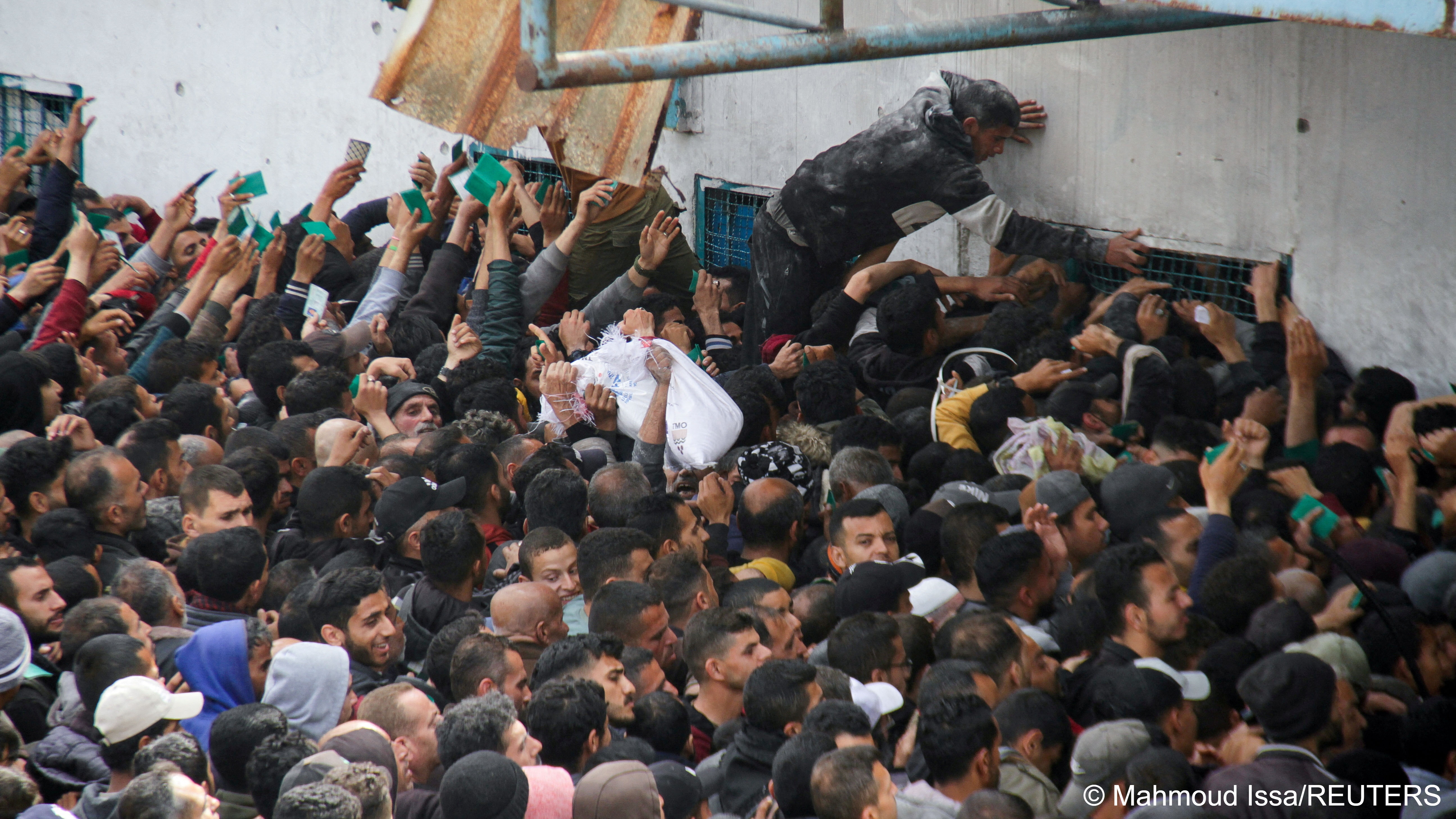 Palestinians, some holding green cards in the air, crowd around an UNRWA warehouse in Gaza waiting for aid
