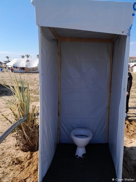 A toilet can be seen in a rudimentary cubicle made of wooden slats and plastic sheeting. In the background are tents