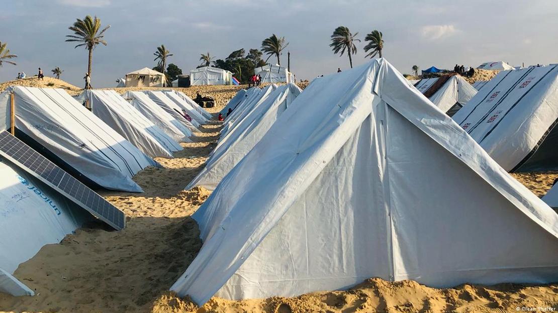 Basic white tents made using plastic sheeting from UNICEF are lined up close to each other. There are more, larger tents and palm trees in the background