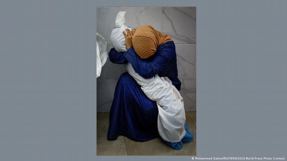 A woman in blue dress and a yellow headscarf cradles the body of a dead child that has been wrapped in white burial material 
