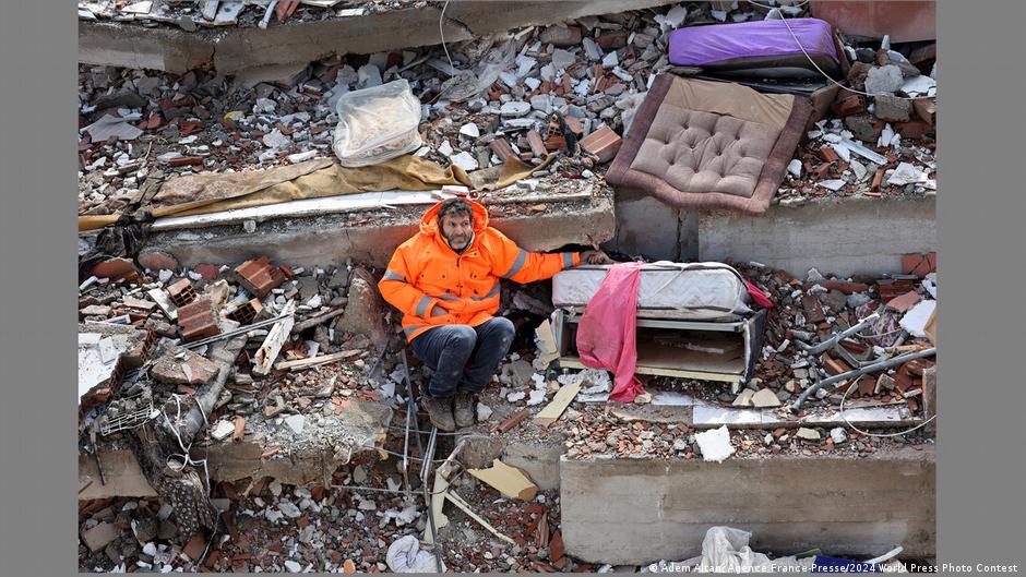 A man in a bright orange jacket sits amidst rubble and debris holding the hand of a corpse that is not visible