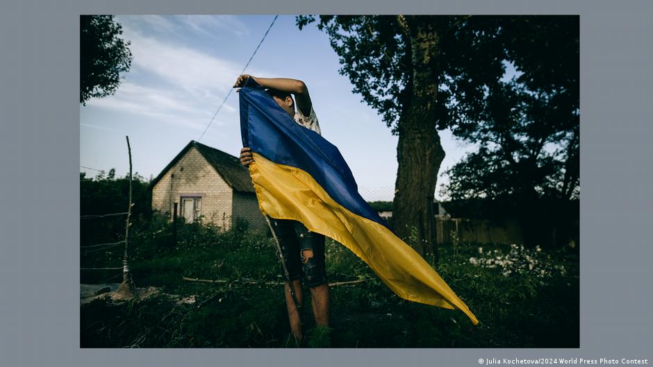 A man in tattered clothes holds up a Ukrainian flag, his face hidden by the flag