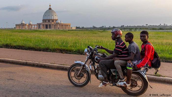 Three young men on a motorbike ride past the Basilica of Our Lady of Peace (photo: DW/E. Lafforgue)