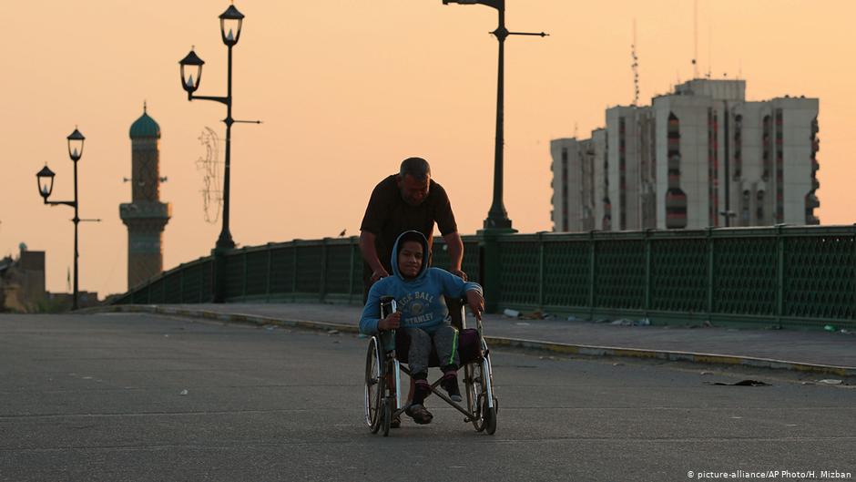 One man pushes another in a wheelchair down a street in Iraq (photo: picture-alliance/AP Photo/H. Mizban)