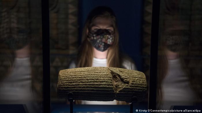 Eine Museumsmitarbeiterin mit medizinischer Maske betrachtet einen beschrifteten Zylinder aus dem Jahr 539 v. Chr.