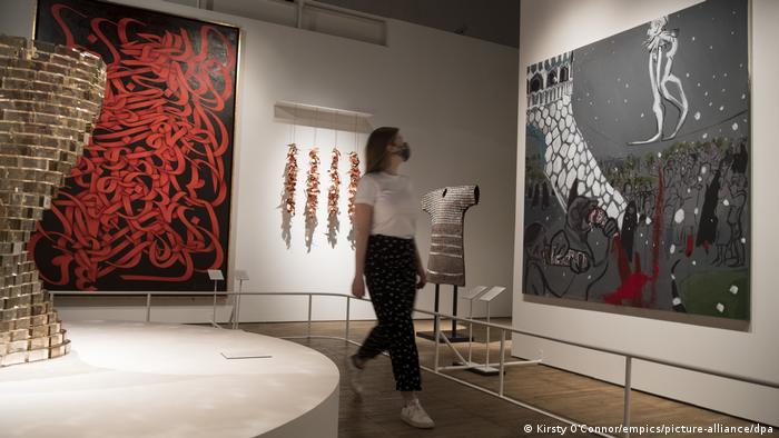 Eine Museumsmitarbeiterin mit medizinischer Maske läuft zwischen mehreren Kunstwerken hindurch, darunter zwei Gemälde, eines rot-schwarz, das andere weiß-grau-schwarz-rot.
