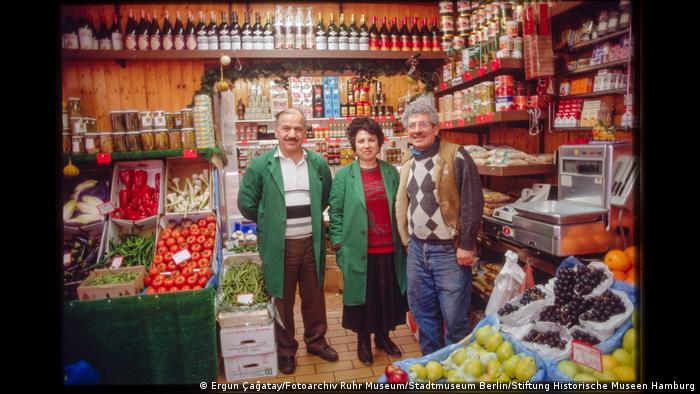 Drei Menschen stehen in einem Lebensmittelladen und lächeln in die Kamera. Aus der Ausstellung "Wir sind von hier. Deutsch-Türkisches Leben 1990".