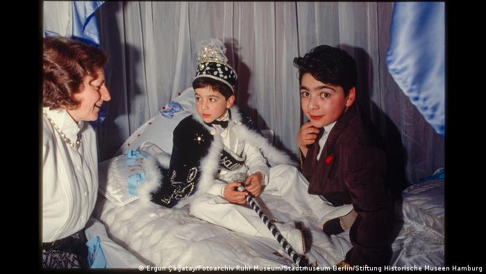 Junge bei seinem Beschneidungsfest in festlicher Kleidung, neben ihm ein anderer Junge und eine Frau. Aus der Ausstellung "Wir sind von hier. Deutsch-Türkisches Leben 1990".