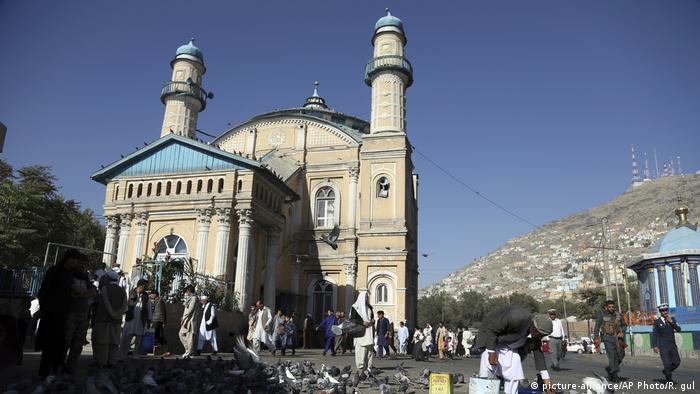 A classical-looking mosque against a backdrop of mountains; people walking past