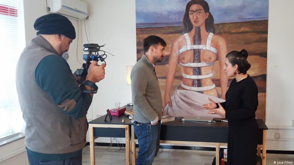 Szene aus dem Film "Ghosts of Afghanistan": Ein Mann spricht mit einer Frau, die von einer dritten Person gefilmt wird; im Hintergrund ein Selbstporträt von Frida Kahlo mit ihren Brüsten