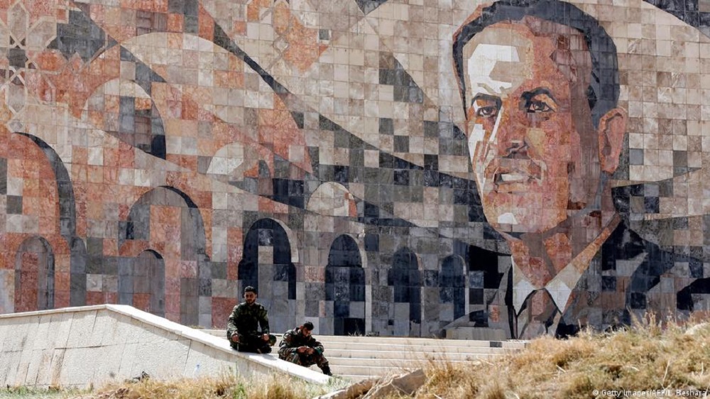  عنصران من القوات السورية يجلسان قرب فسيفساء تمثل صورة الرئيس السوري الراحل حافظ الاسد عند مدخل حرستا قرب دمشق في 25 آذار/مارس 2018.  
