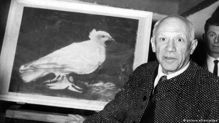 Pablo Picasso steht neben seinem Gemälde einer Friedenstaube.