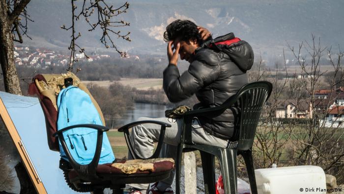Ein Mann sitzt auf einem Plastikstuhl, im Hintergrund sind ein Fluss oder See und Berge zu sehen