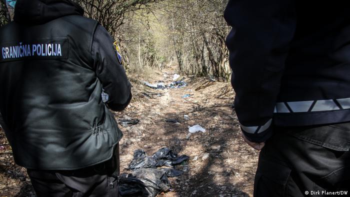  Zwei Personen stehen mit dem Rücken zur Kamera, auf dem Rücken der Jacke der linken steht Grenzpolizie auf bosnisch/kroatisch/serbisch, vor den beiden ist ein Waldweg zu sehen, auf dem Abfall liegt