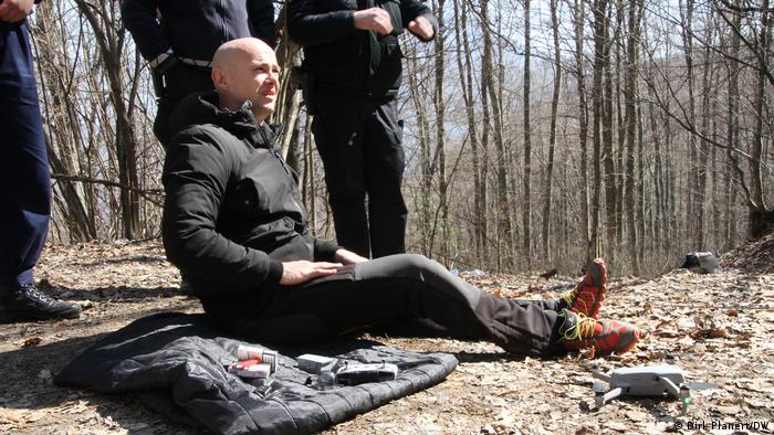  Ein Mann sitzt auf dem Waldboden, neben ihm liegt eine Jacke, dahinter stehen drei weitere Personen