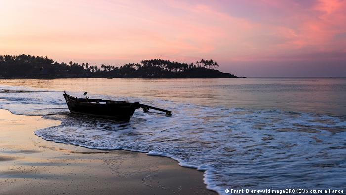 Ein Strand im pinken Sonnenuntergang ist zu sehen. Ein Holzboot liegt im seichten Wasser und im Hintergrund sind die Umrisse von Bäumen zu sehen.