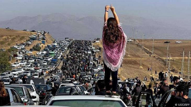 A young woman without a headscarf and with her back to the camera stands on the roof of a car with her hands in the air looking at a long line of people and cars on a motorway (photo: UGC/AFP)