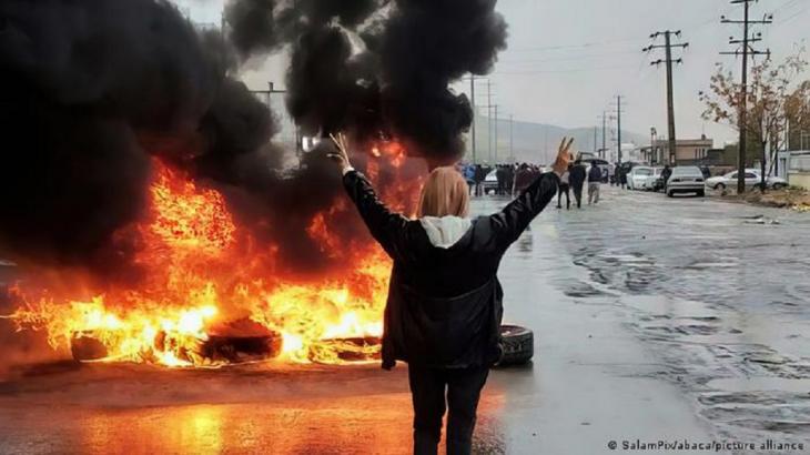 A woman with her back to the camera holds up her hands and makes the victory sign in front of burning tyres (photo: SalamPix/abaca/picture alliance)