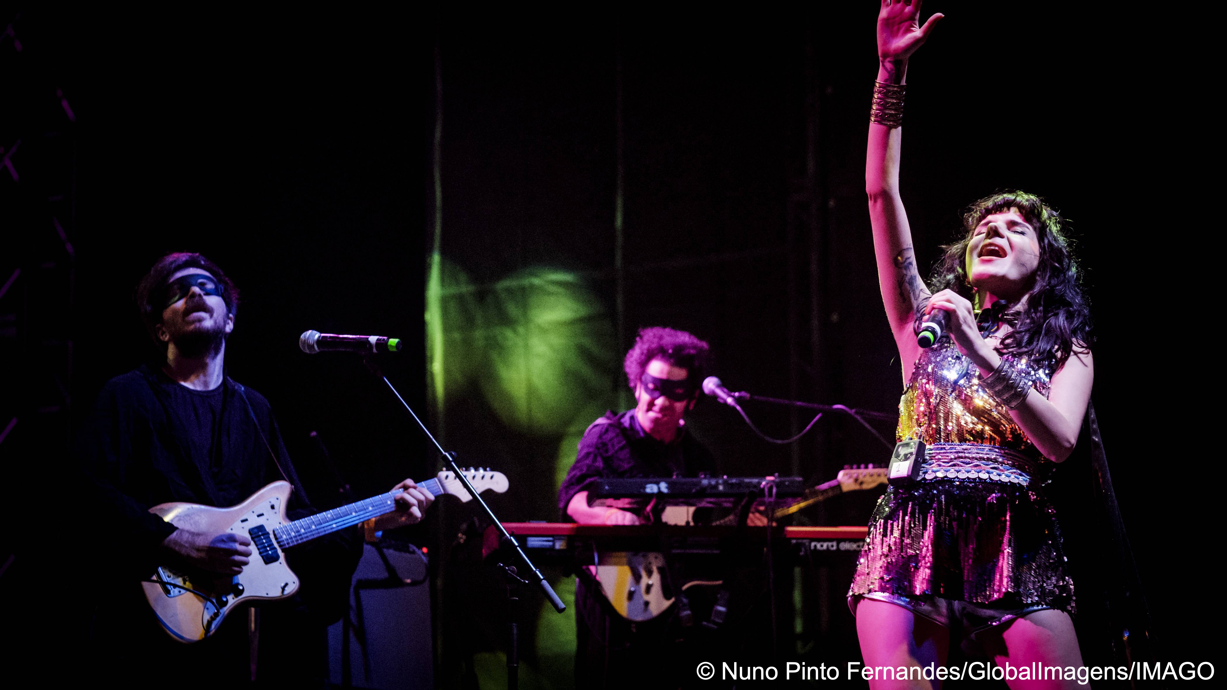 Gaye Su Akyol stretches her right arm into the air while singing on stage alongside two masked musicians (photo: Nuno Pinto Fernandes/GlobalImagens/IMAGO)